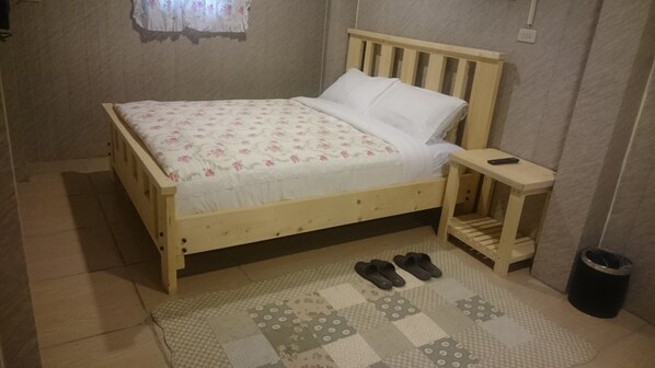 Una cama de madera con colcha estampada y almohadas blancas, una mesita de noche con un control remoto, y un par de pantuflas sobre una alfombra con diseño.