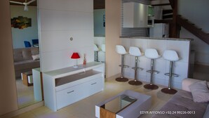 A modern kitchen with white cabinets, a red lamp, and a bar area with white stools. - Residência Angra Deep Blue (Angra dos Reis)