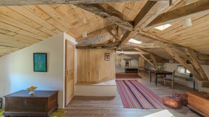 Een zolderkamer met zichtbare houten balken, een houten tafel, een bankje en een kleurrijk gestreept tapijt.