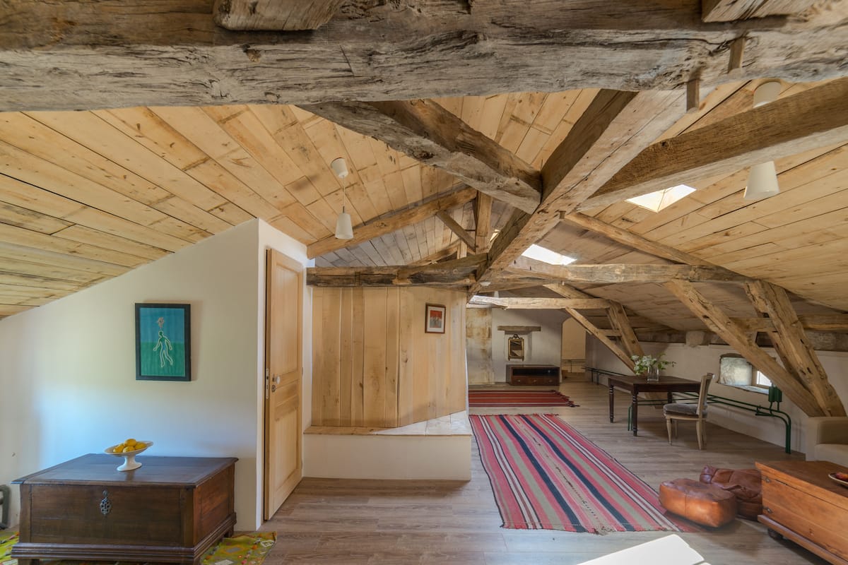 Un dormitorio en el ático con vigas de madera a la vista, una mesa de madera, un banco y una alfombra colorida a rayas.