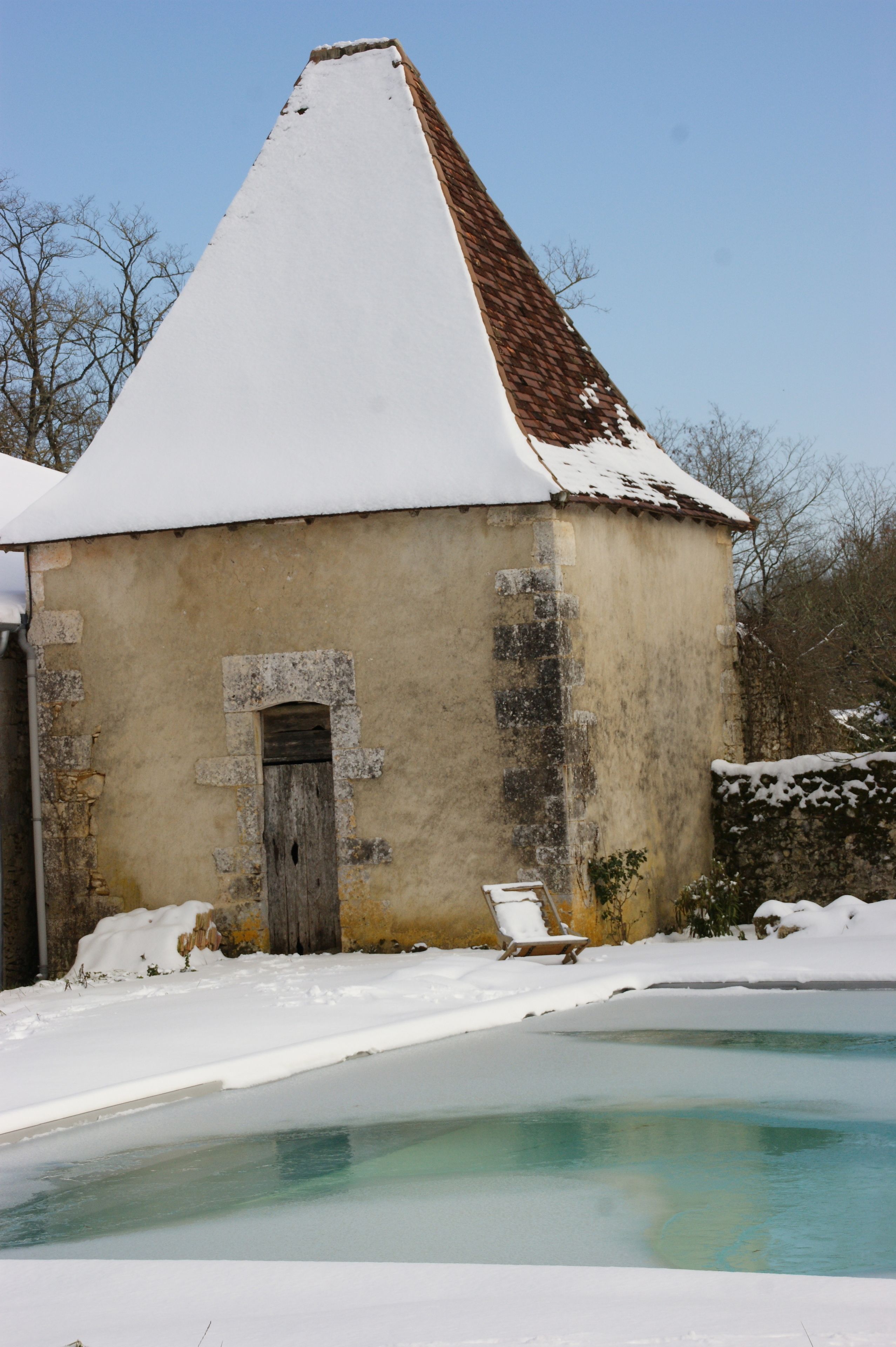 A small, rustic building with a snow-covered roof and a wooden door, adjacent to a partially frozen pool.