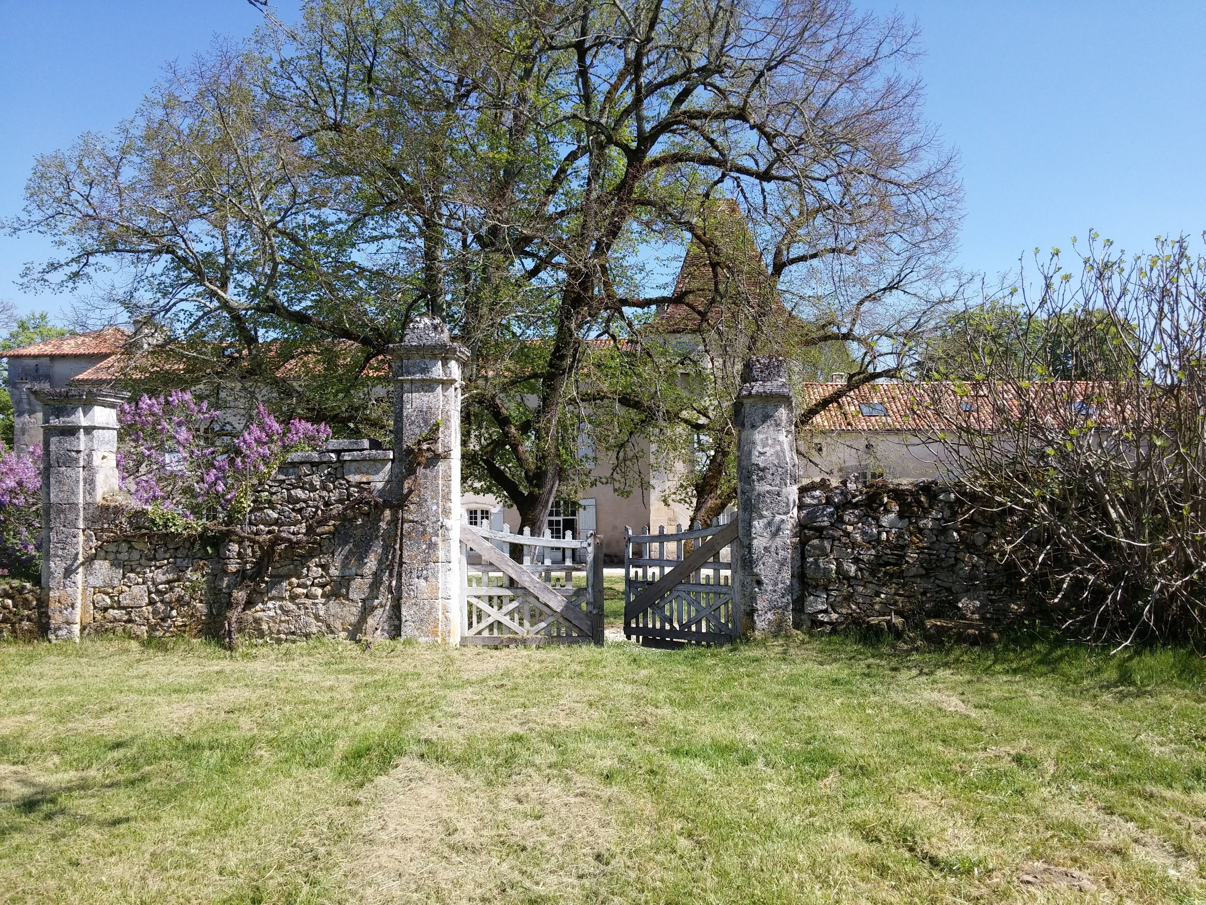 Een stenen poort met houten omheining, omringd door gras en bomen, die leidt naar een gebouw met pannendak.