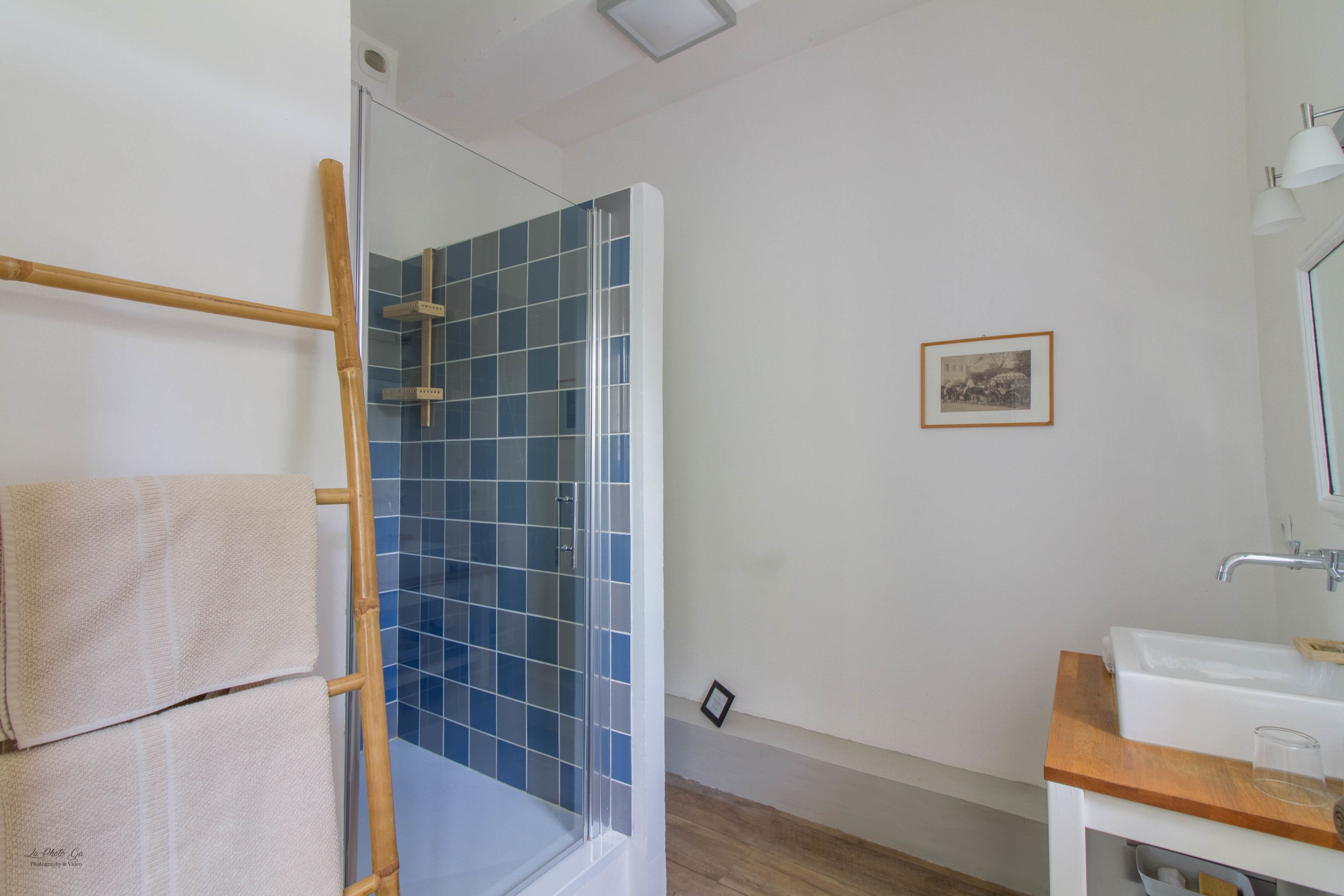 Un baño con cabina de ducha de vidrio, un perchero de madera, un lavamanos con espejo y un cuadro en la pared.