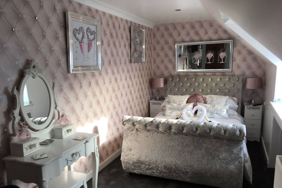 A bedroom with a tufted headboard, a large mirror, a dresser, and a bed with decorative pillows and a heart-shaped towel arrangement.