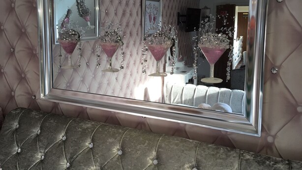 A mirror with a metallic frame reflecting a tufted wall and a tufted bench. - Gleninver Guest House (Inverness)