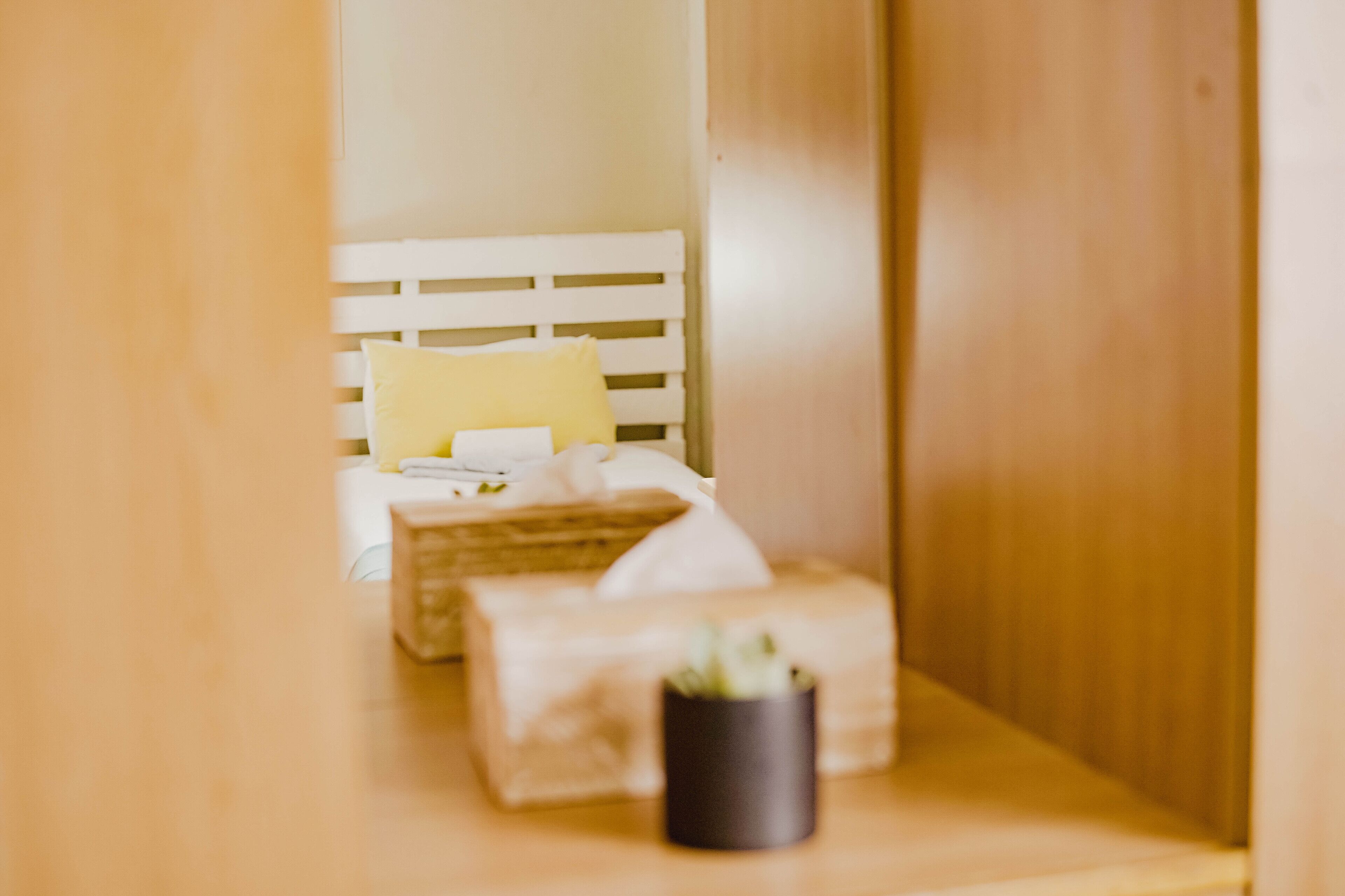 A narrow hallway leading to a bed with a white headboard, a yellow pillow, and a small potted plant on a wooden nightstand.