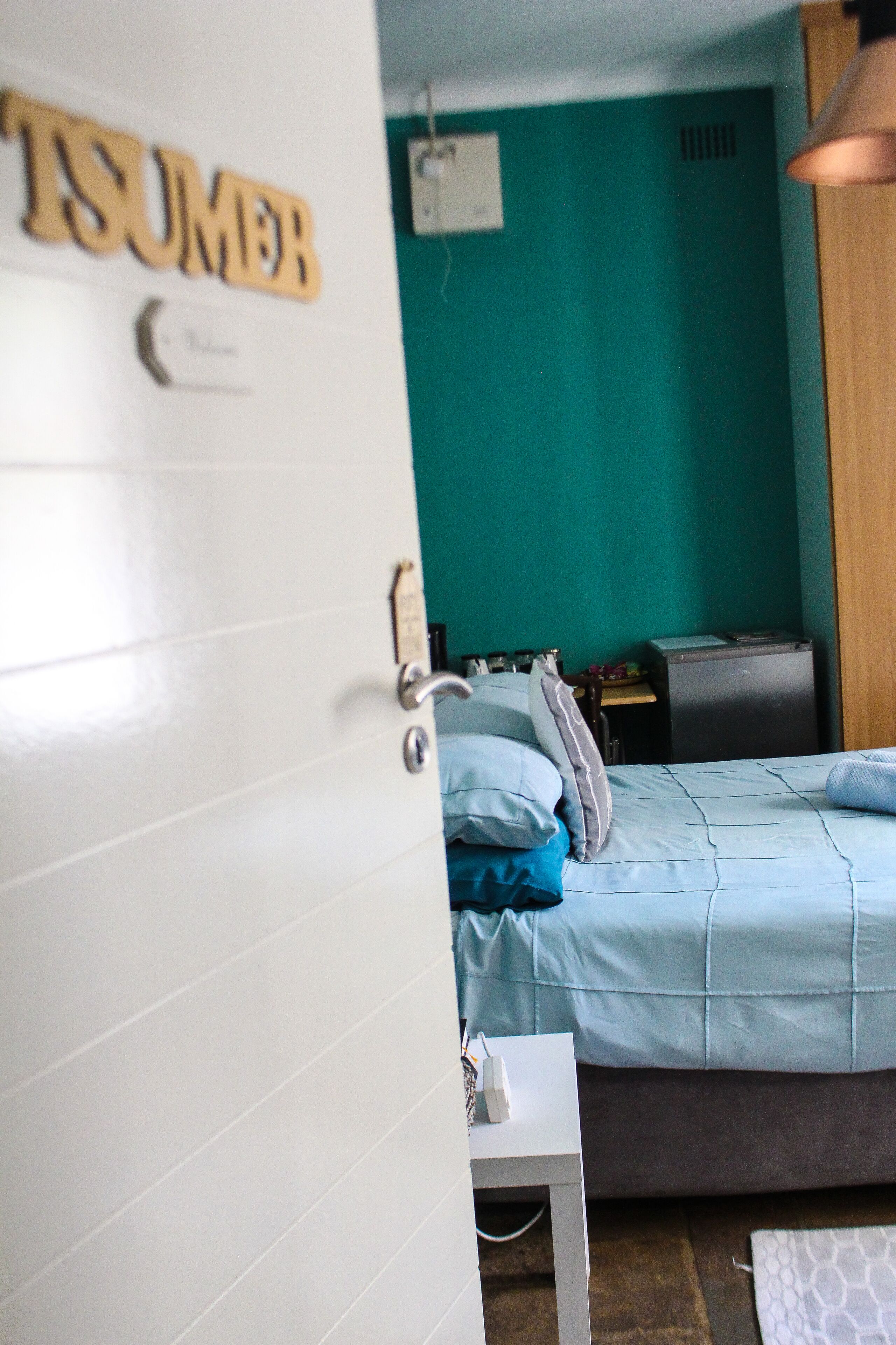 A room with a teal wall, a bed with blue bedding, a small white table, and a wooden cabinet.