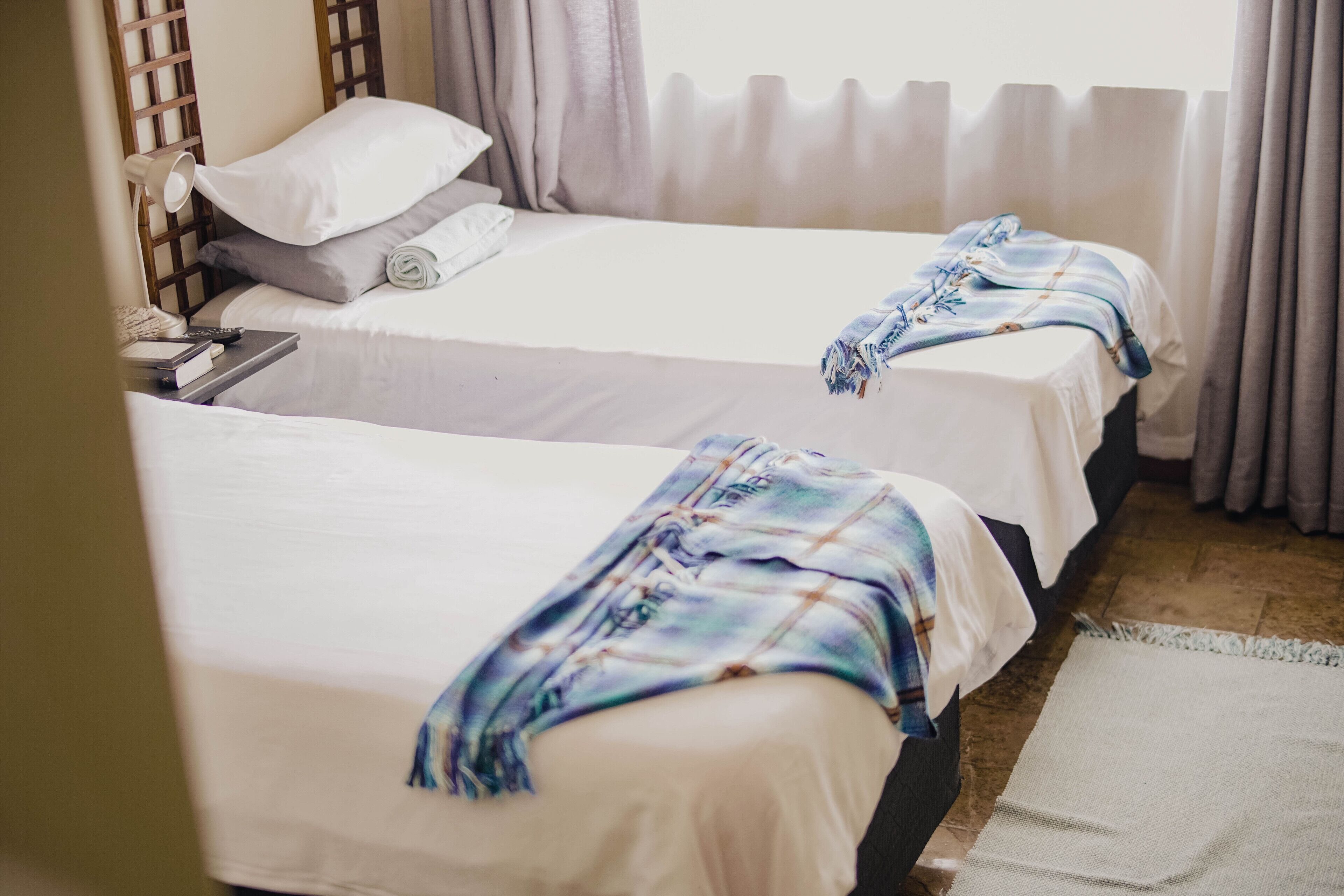 Two single beds with white linens, a bedside table with a lamp, and a window with curtains.