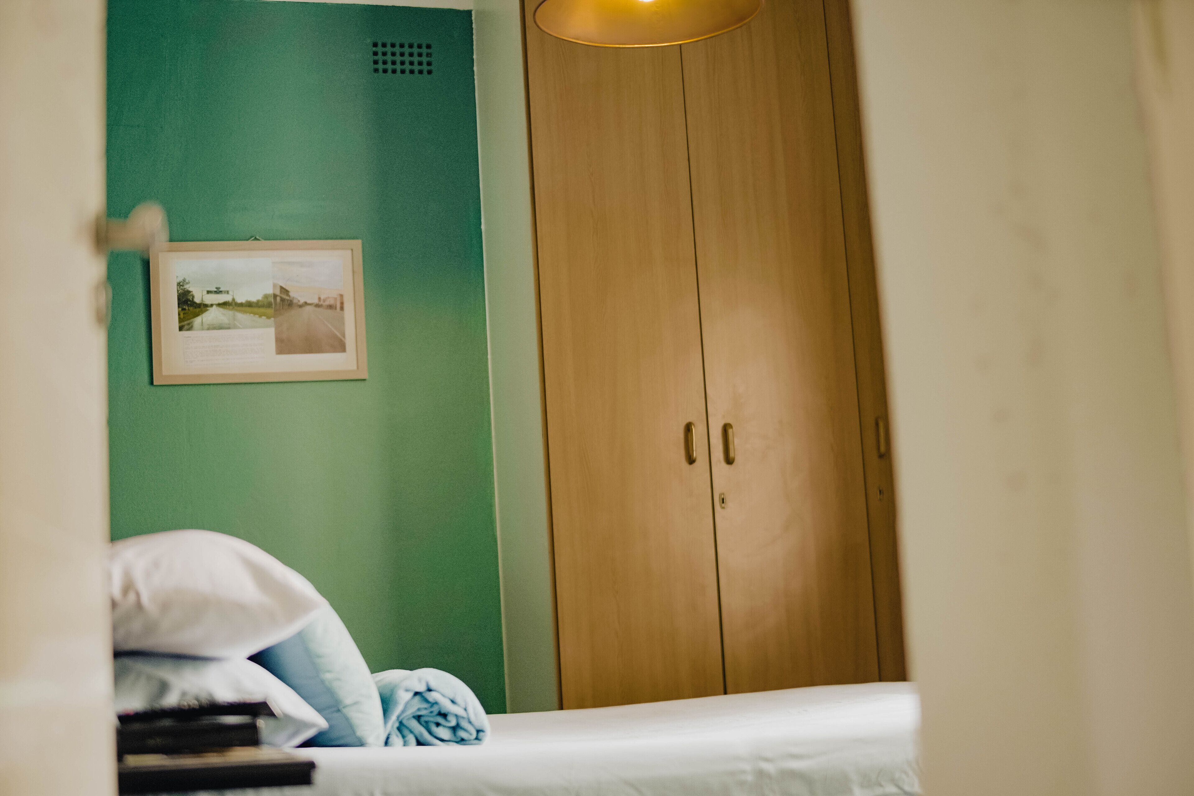 A room with a bed, a wooden wardrobe, a framed picture on the wall, and a lamp.