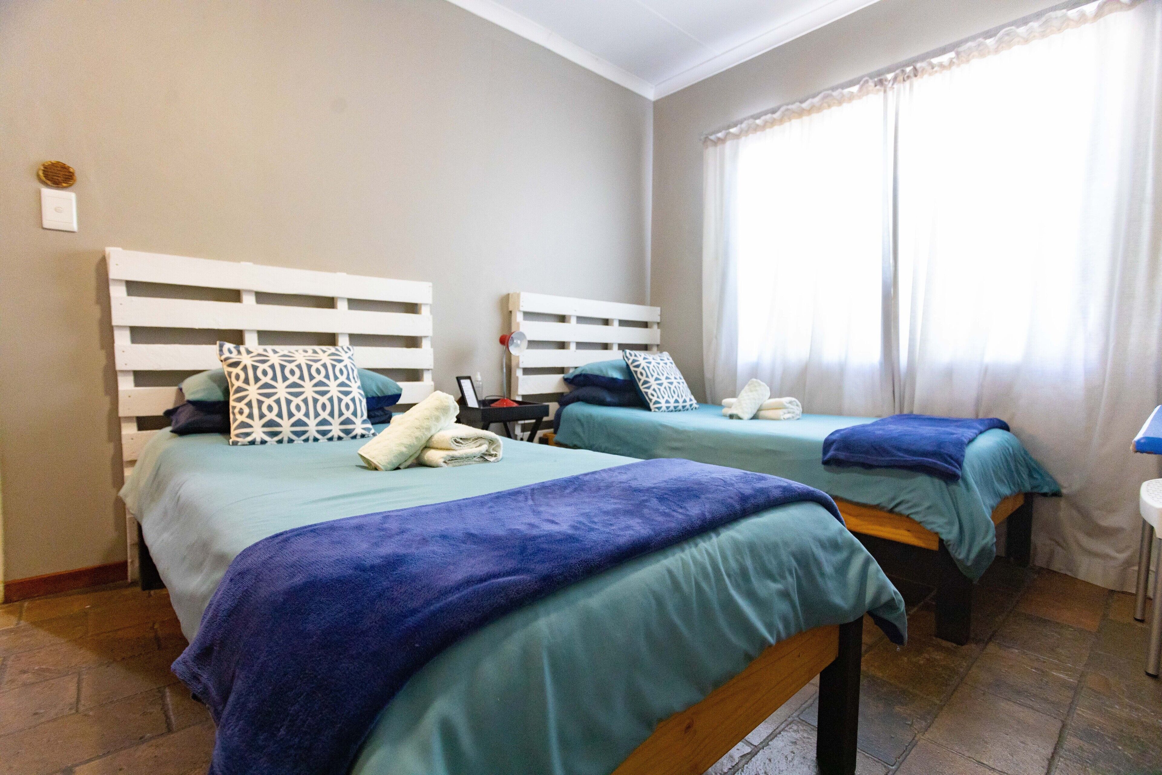 A room with two beds, each with a headboard and pillows, a bedside table with a lamp, and a window with curtains.