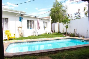 A swimming pool with a tiled edge, surrounded by a grassy yard and a white building with windows and a door. - Kate's Nest Guesthouse (Windhoek)
