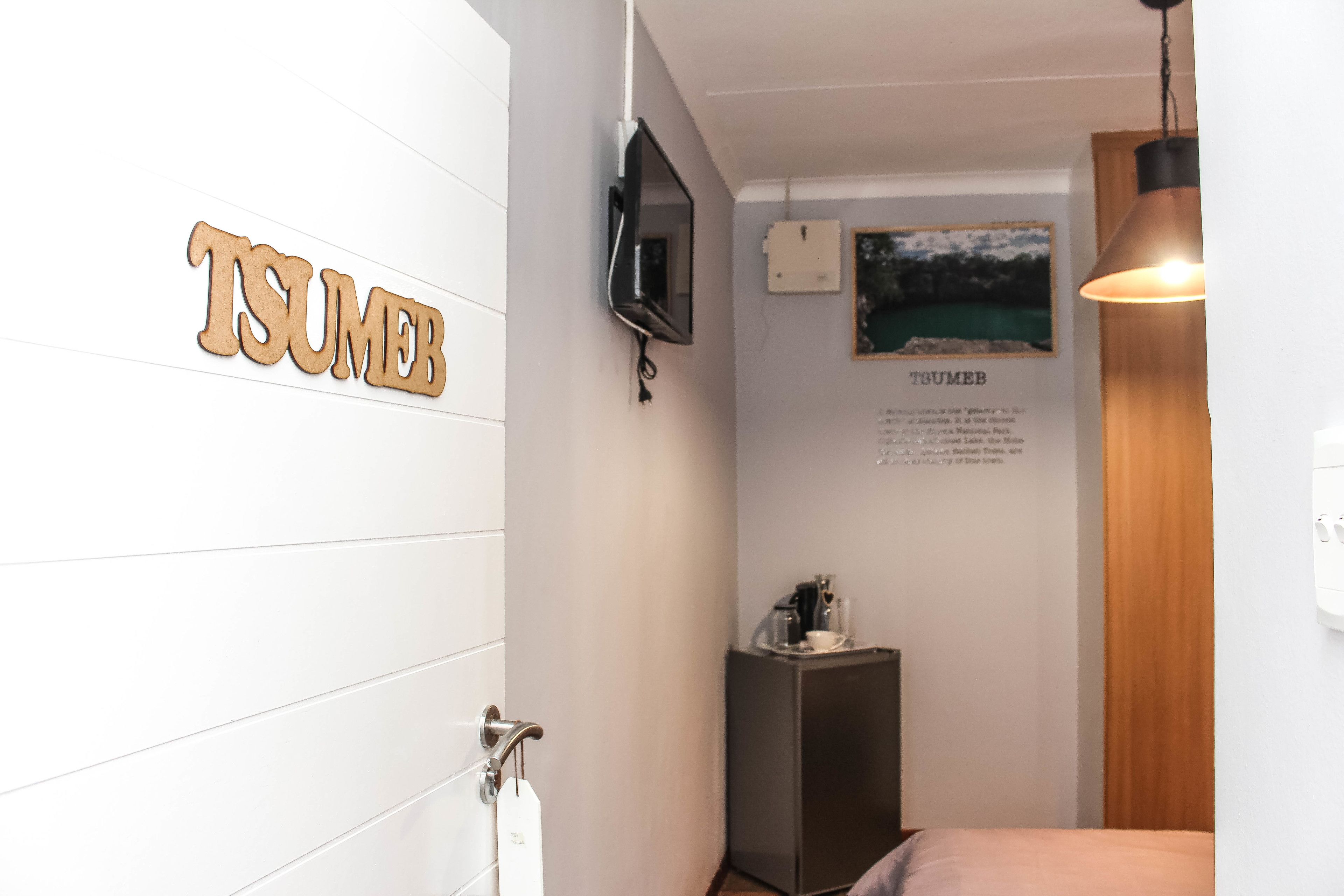 A hotel room with a wooden sign reading "TSUMEB," a mounted television, a small table with a coffee maker, and a hanging light fixture.