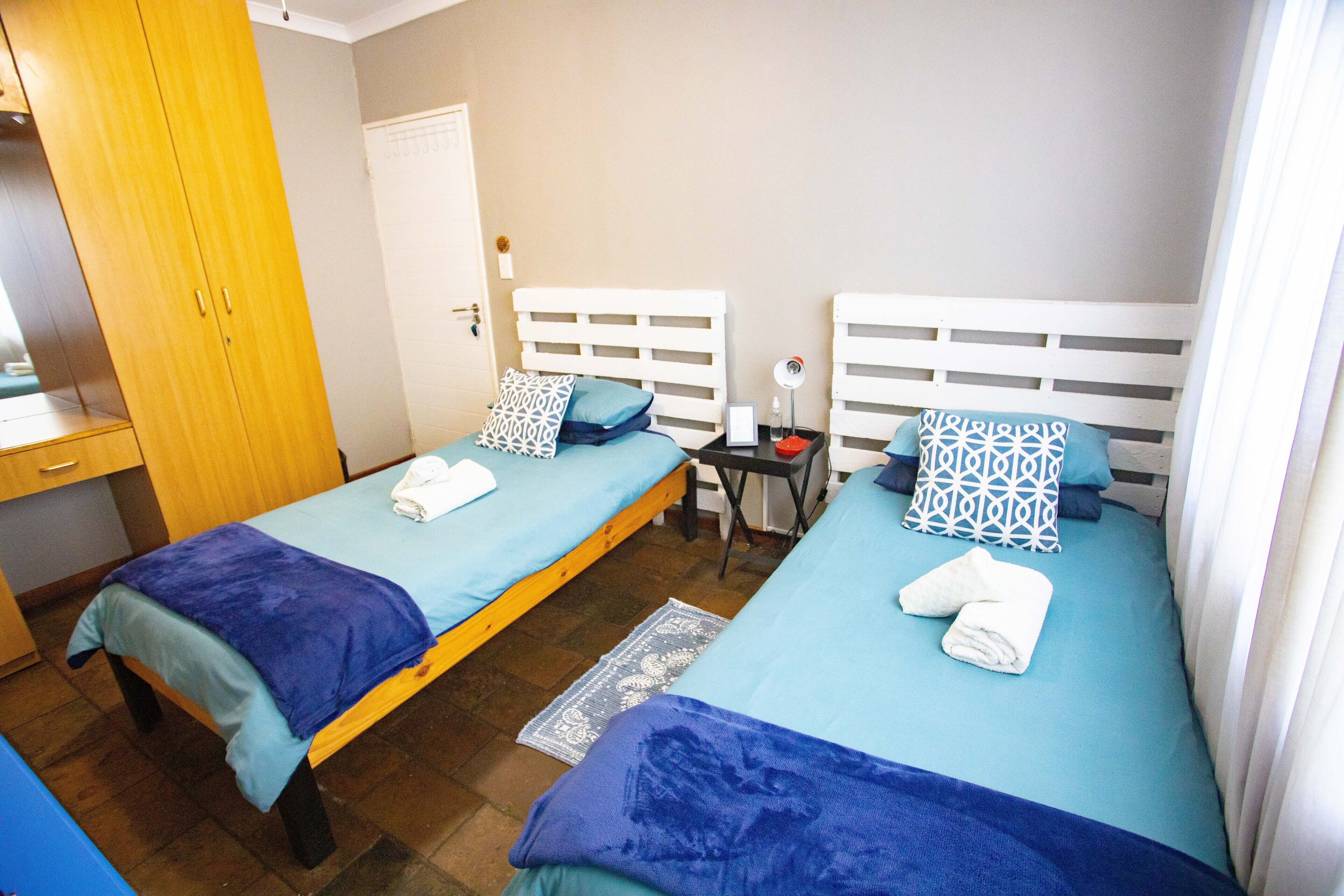 Een kamer met twee enkele bedden, elk voorzien van een blauwe sprei en gekleurde kussens, een houten garderobe en een tafeltje met een spiegel en fles.
