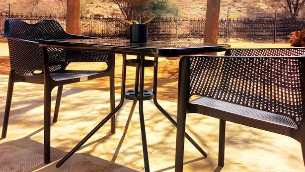 An outdoor seating area with a metal table and chairs, surrounded by trees and a fence. - Kate's Nest Guesthouse (Windhoek)