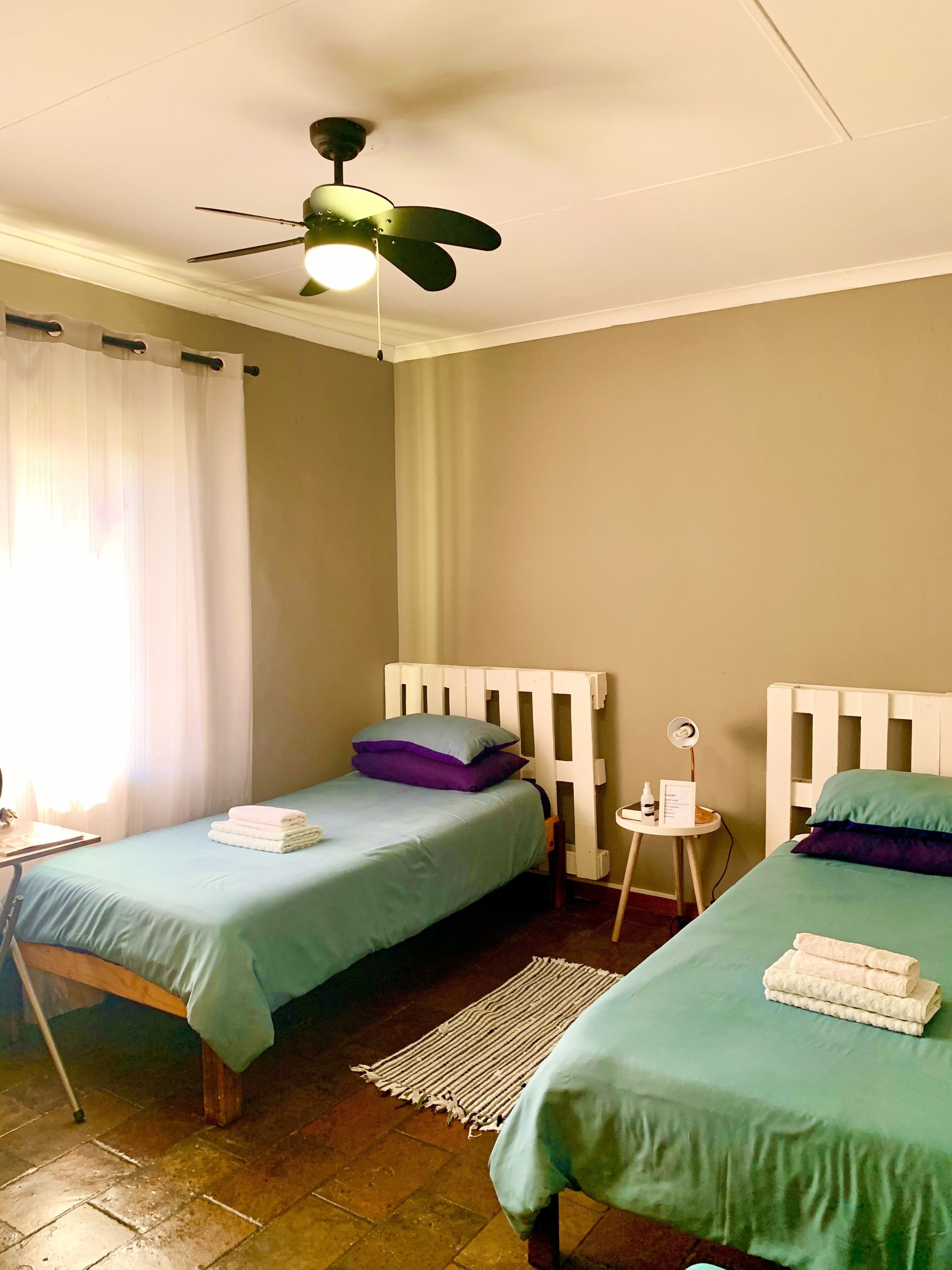 Una habitación con dos camas individuales, un ventilador de techo, una mesita con lámpara y una ventana con cortinas.