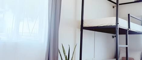 A bunk bed with a black frame and white bedding, a potted plant, a window with curtains, and a small table.