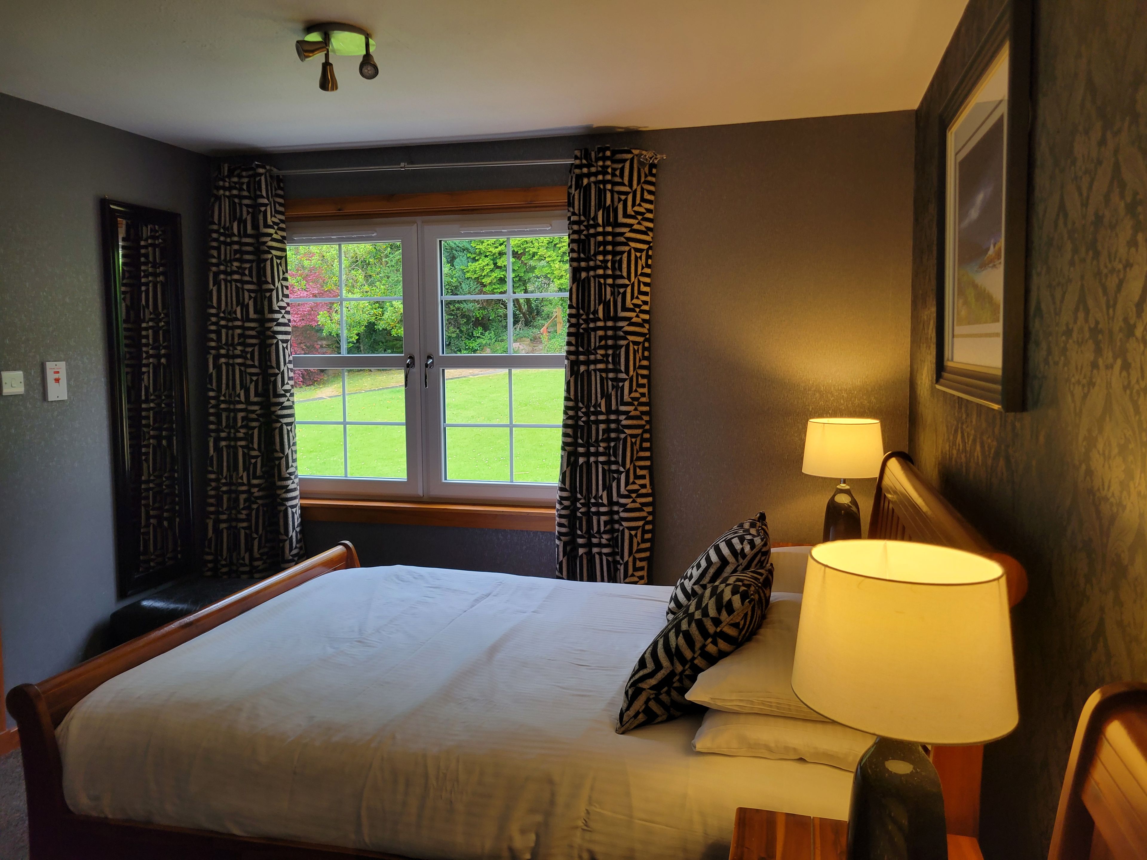 Un dormitorio con cama, lámpara de noche, ventana con cortinas y vista a un jardín.