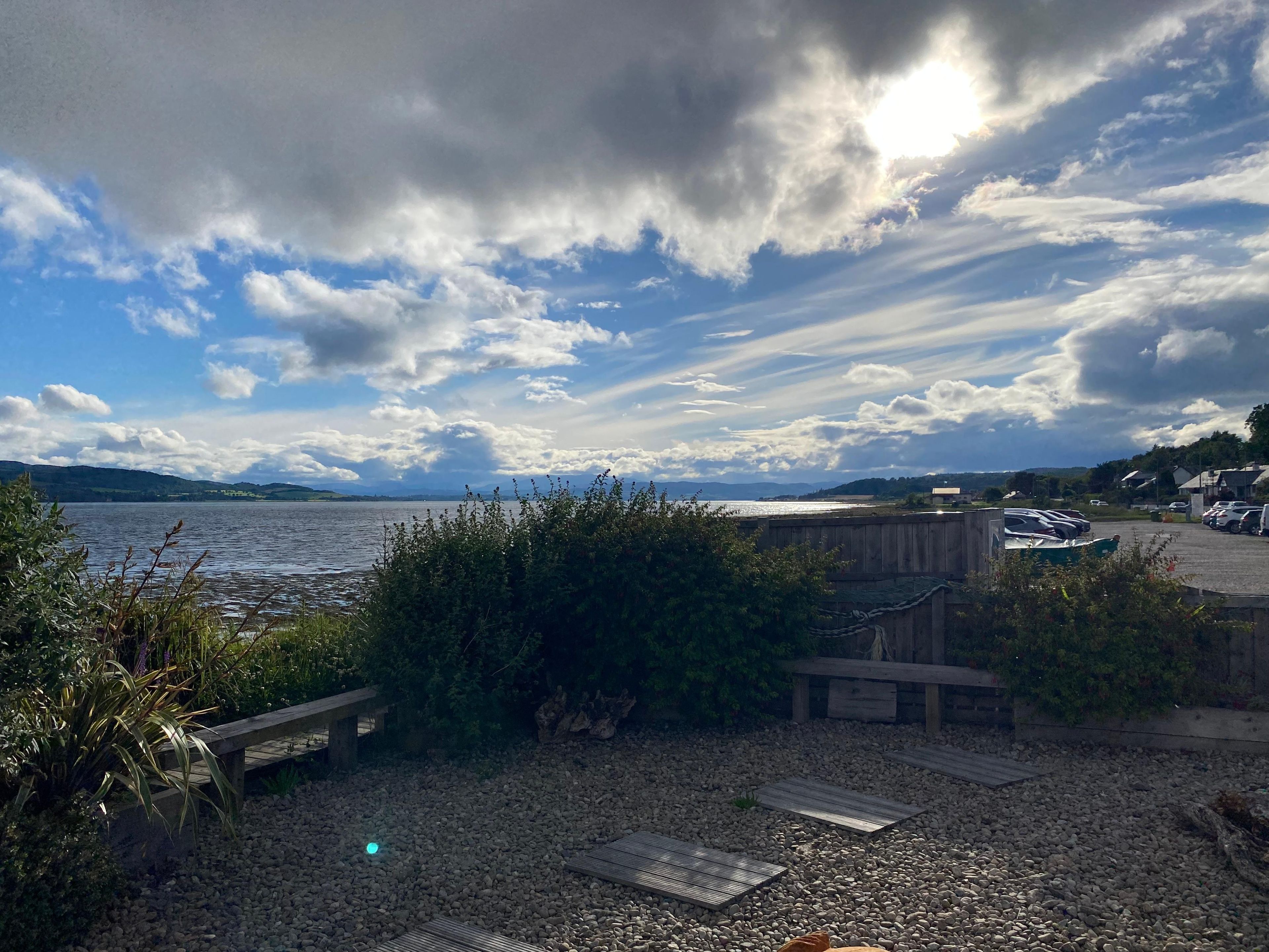 A lakeside view with a wooden deck, pebbles, and a parking area.