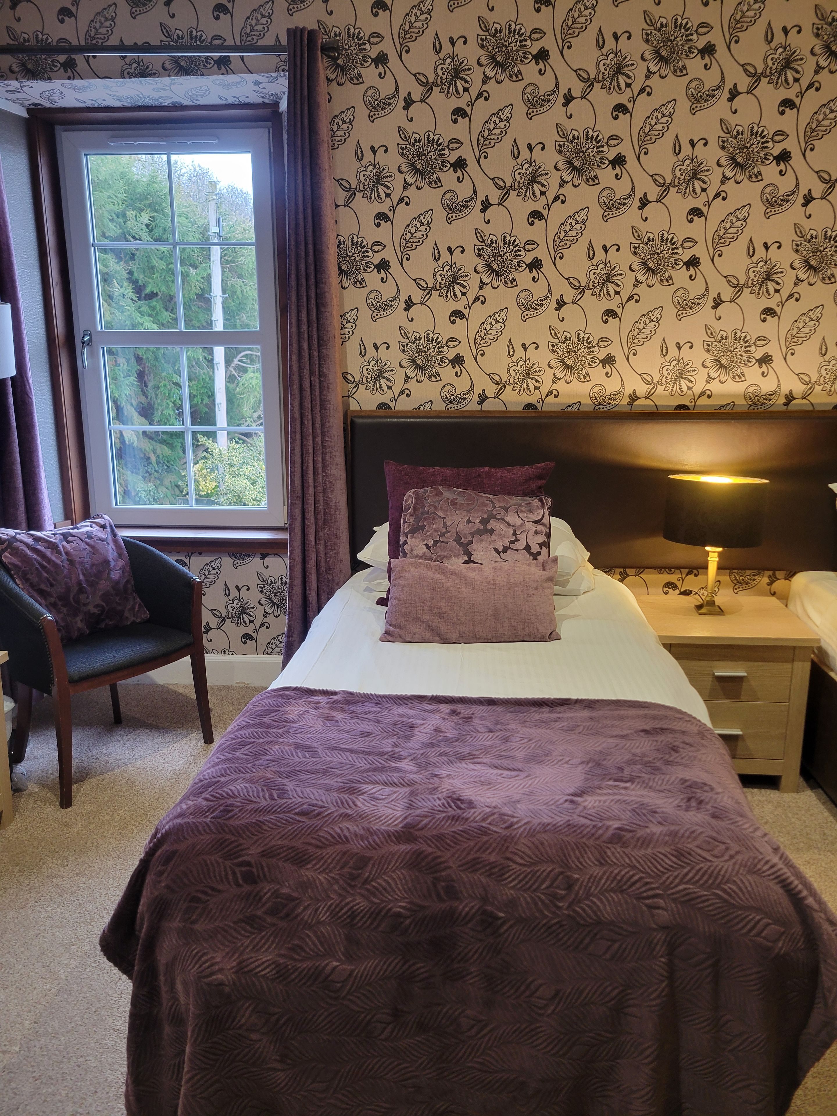 Een slaapkamer met een bed, een stoel, een nachtkastje en een raam met gordijnen.