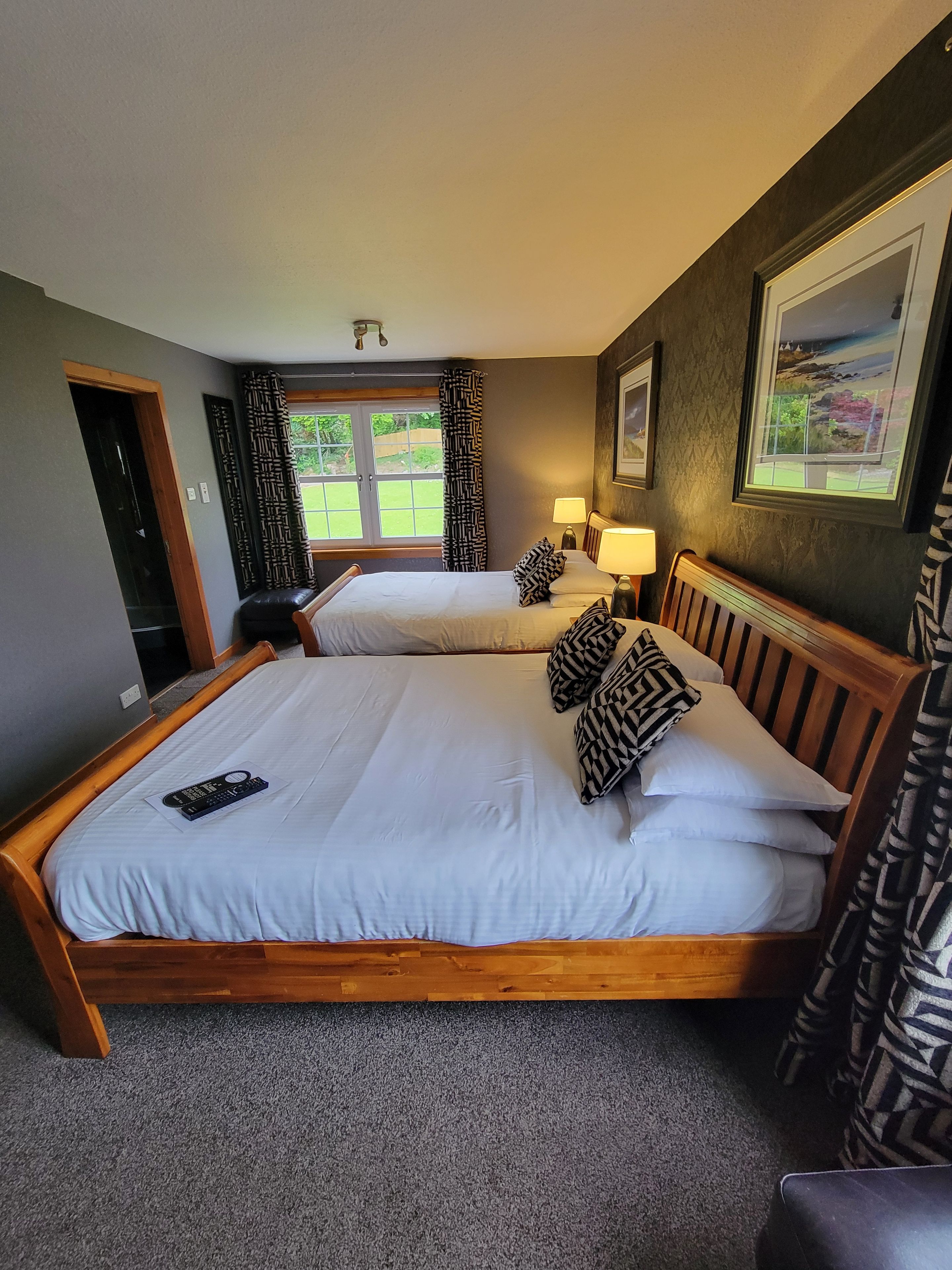 A double bed with a remote control, two bedside lamps, patterned pillows, and a view of a garden through a window.