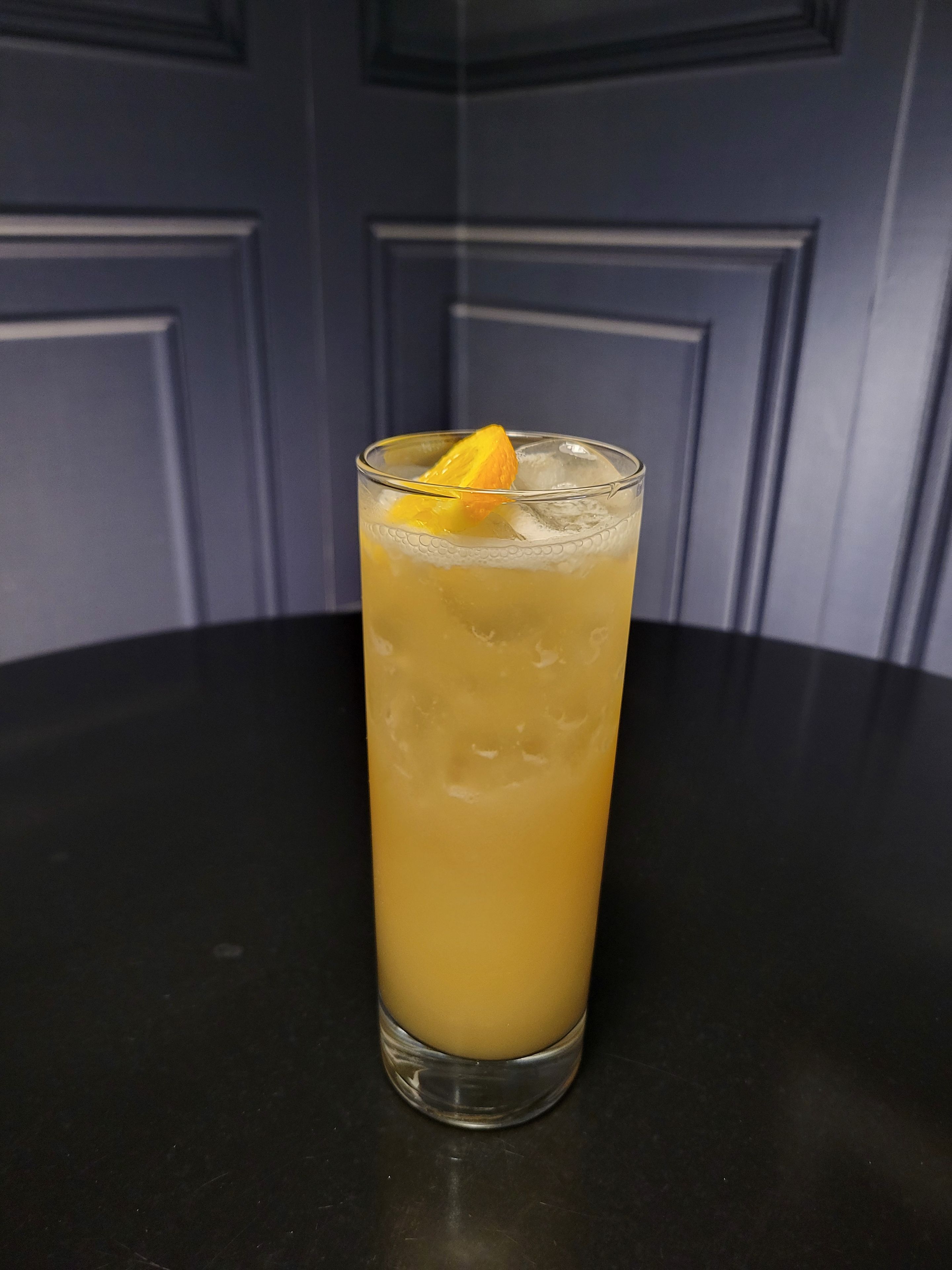 A tall glass with a yellow beverage, ice cubes, and a lemon slice on a dark surface.