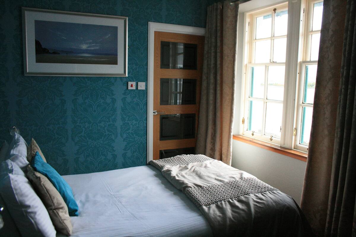 A bedroom with a bed, a door, a window, and a framed picture on the wall.