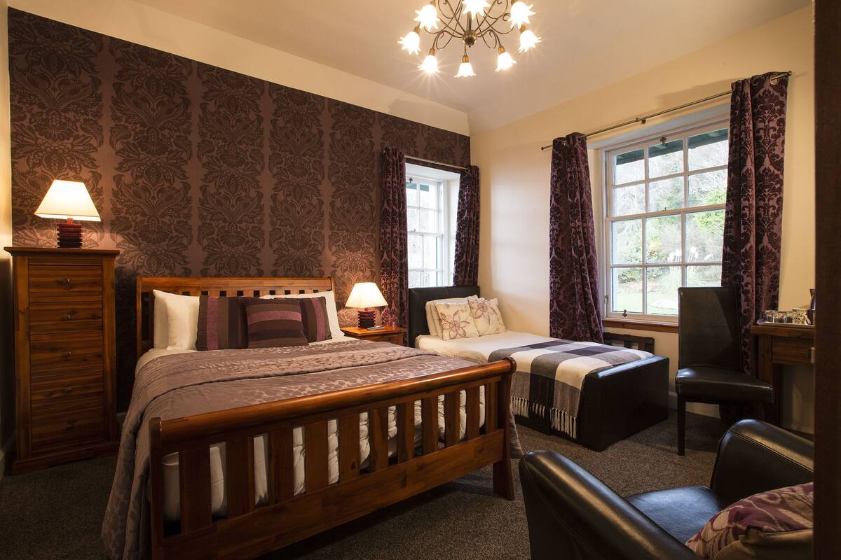 A bedroom with a wooden bed, a nightstand with a lamp, a chair, and a window with curtains.