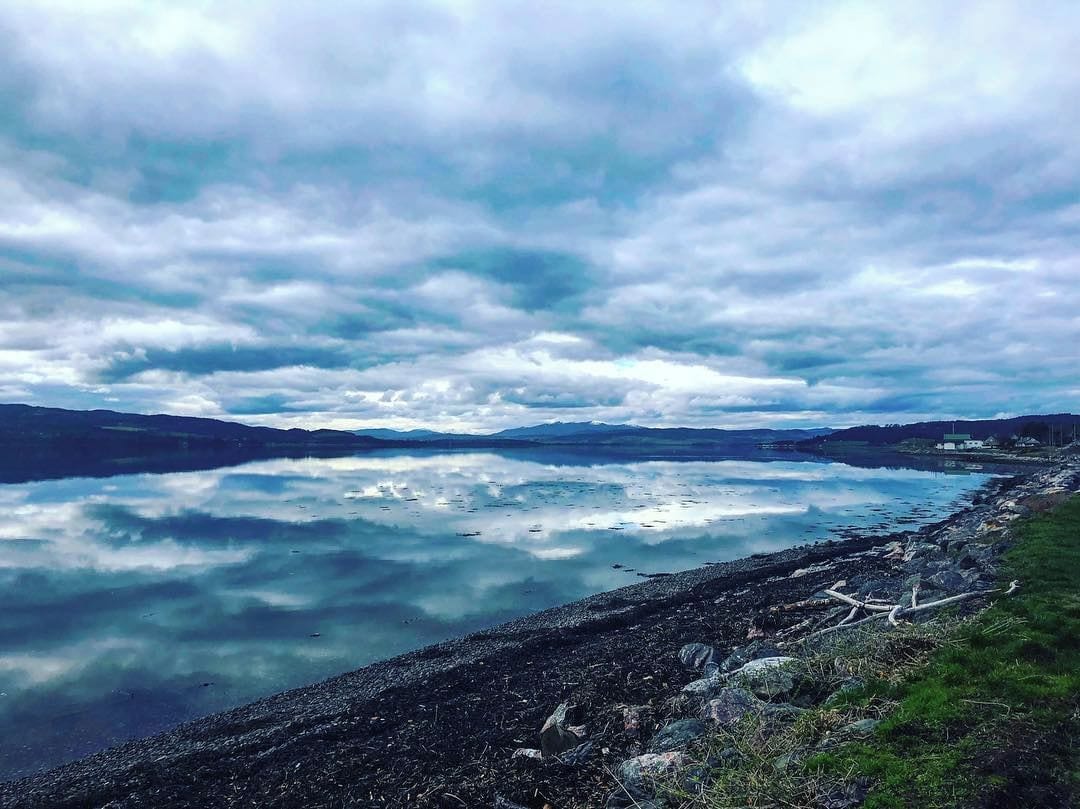 Un llac tranquil amb riba rocosa i muntanyes llunyanes sota un cel nuvolós.