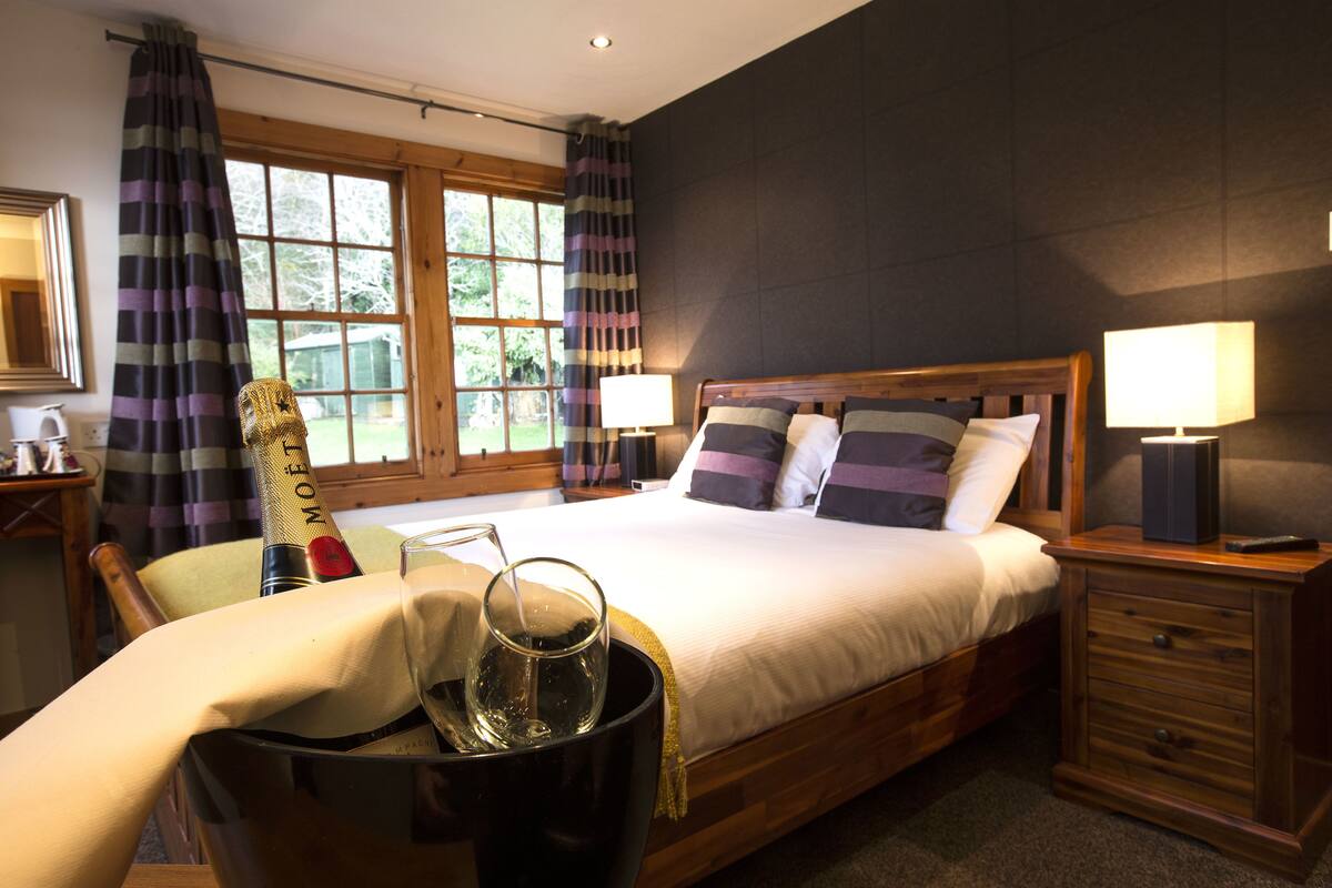 A hotel room with a large bed, two bedside tables with lamps, a wooden nightstand, and a view of the outdoors through large windows.