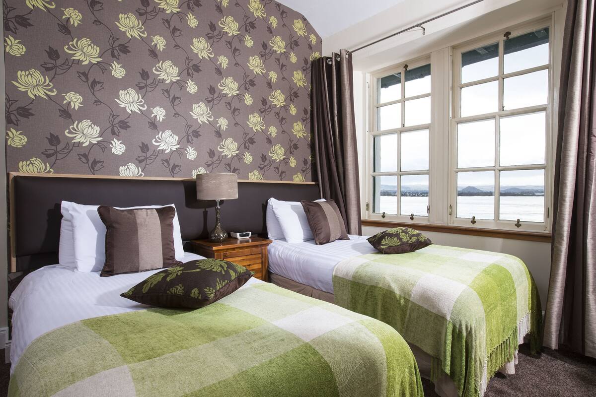 A hotel room with two beds, floral wallpaper, a window with a view, and a nightstand with a lamp.