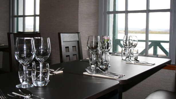 A dining table set with multiple wine glasses, forks, and knives, with a view of a body of water through large windows. - North Kessock Hotel (Inverness)