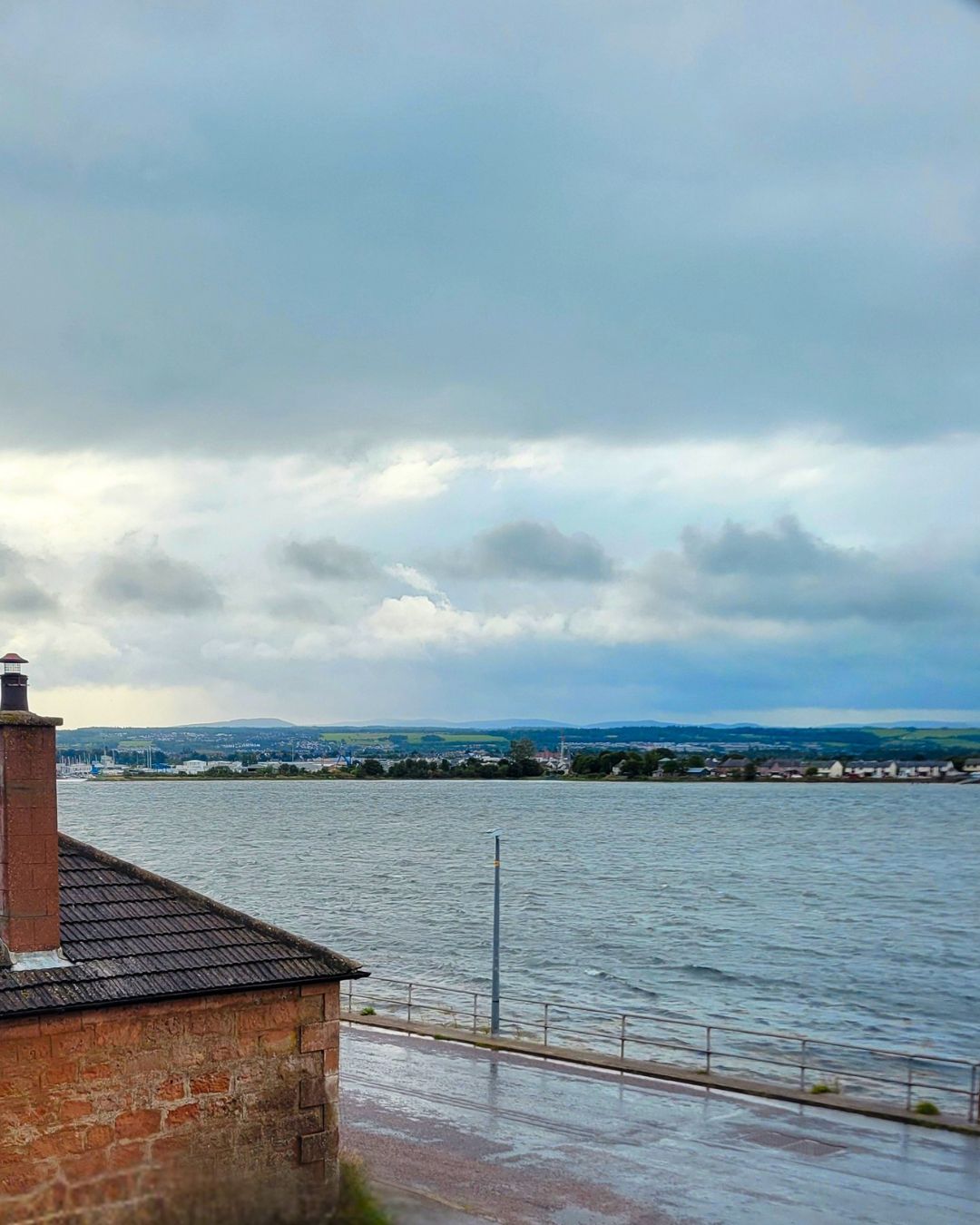 A waterfront view with a brick building in the foreground and a distant town across the water.