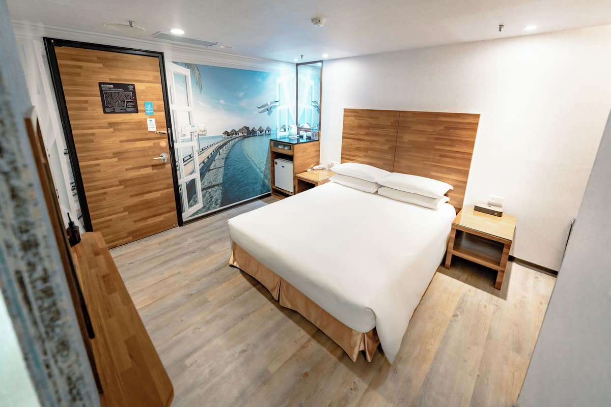 A hotel room with a large bed, wooden headboard, a small desk, and a view of a coastal scene through a glass door.