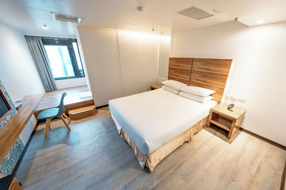 A modern hotel room with a large bed, a desk with a chair, a bedside table, and a window with curtains.