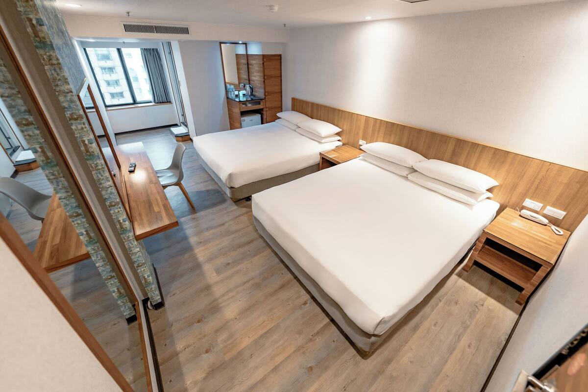 A modern hotel room with two beds, a wooden floor, a small desk, and a window with a view of buildings.