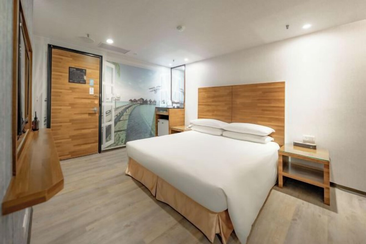 A modern hotel room with a large bed, wooden headboard, a small bedside table, and a wall-mounted TV.