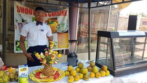 A man standing behind a counter with a blender, a fruit display, and a menu board advertising smoothies. - Glamossay Hotel (Sunyani)