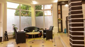 A modern living room with a black leather sofa, a round coffee table, and black armchairs. Large windows provide a view of greenery outside. - Glamossay Hotel (Sunyani)