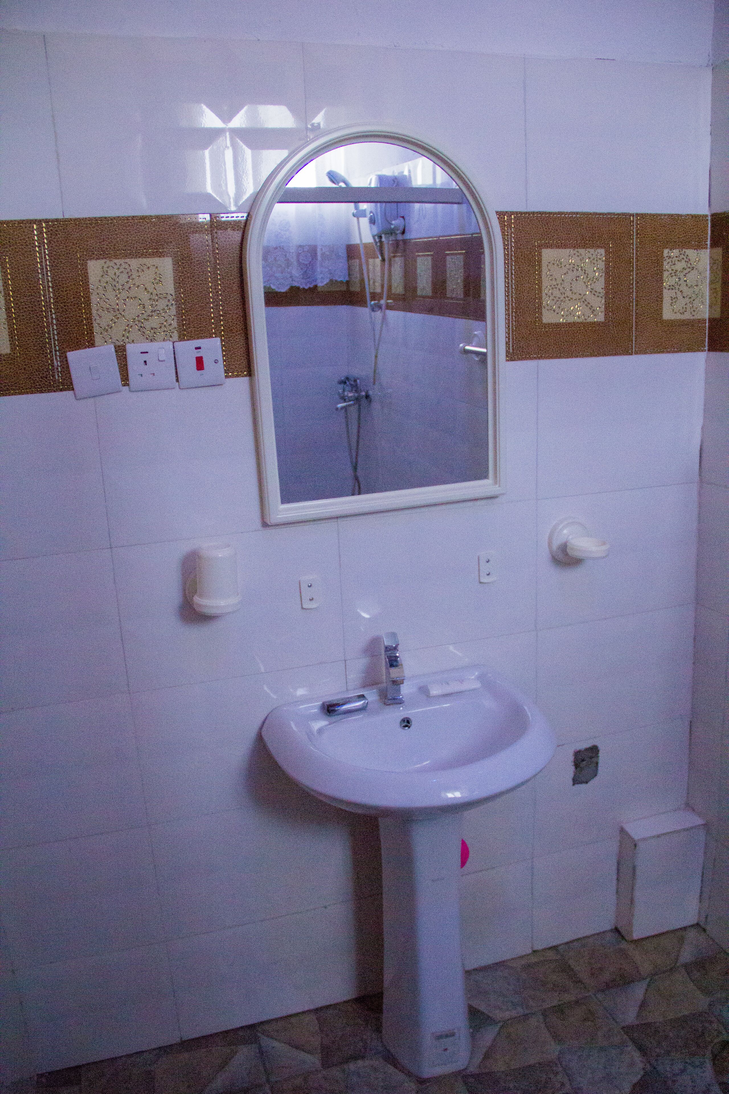 Un baño con un lavamanos blanco, un espejo y una ducha. Hay dispensadores de jabón en la pared y un perchero para toallas.