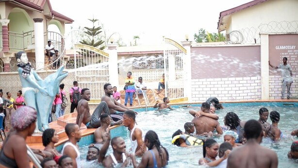 A swimming pool area with people enjoying the water, a statue, and a gated entrance. - Glamossay Hotel (Sunyani)