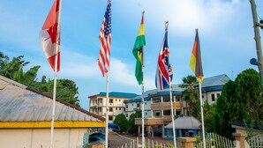 A building with multiple flags, including the American and German flags, and a fenced area with a building in the background. - Glamossay Hotel (Sunyani)