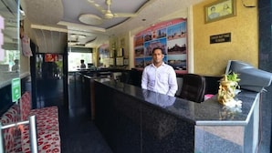 A hotel reception area with a receptionist, a check-in counter, a wall with a welcome poster, and a ceiling fan. - Hotel Mohit Palace (Jaipur)