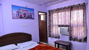 A bedroom with a bed, a wooden headboard, a red blanket, a small table, a wall-mounted air conditioner, and a door. - Hotel Mohit Palace (Jaipur)