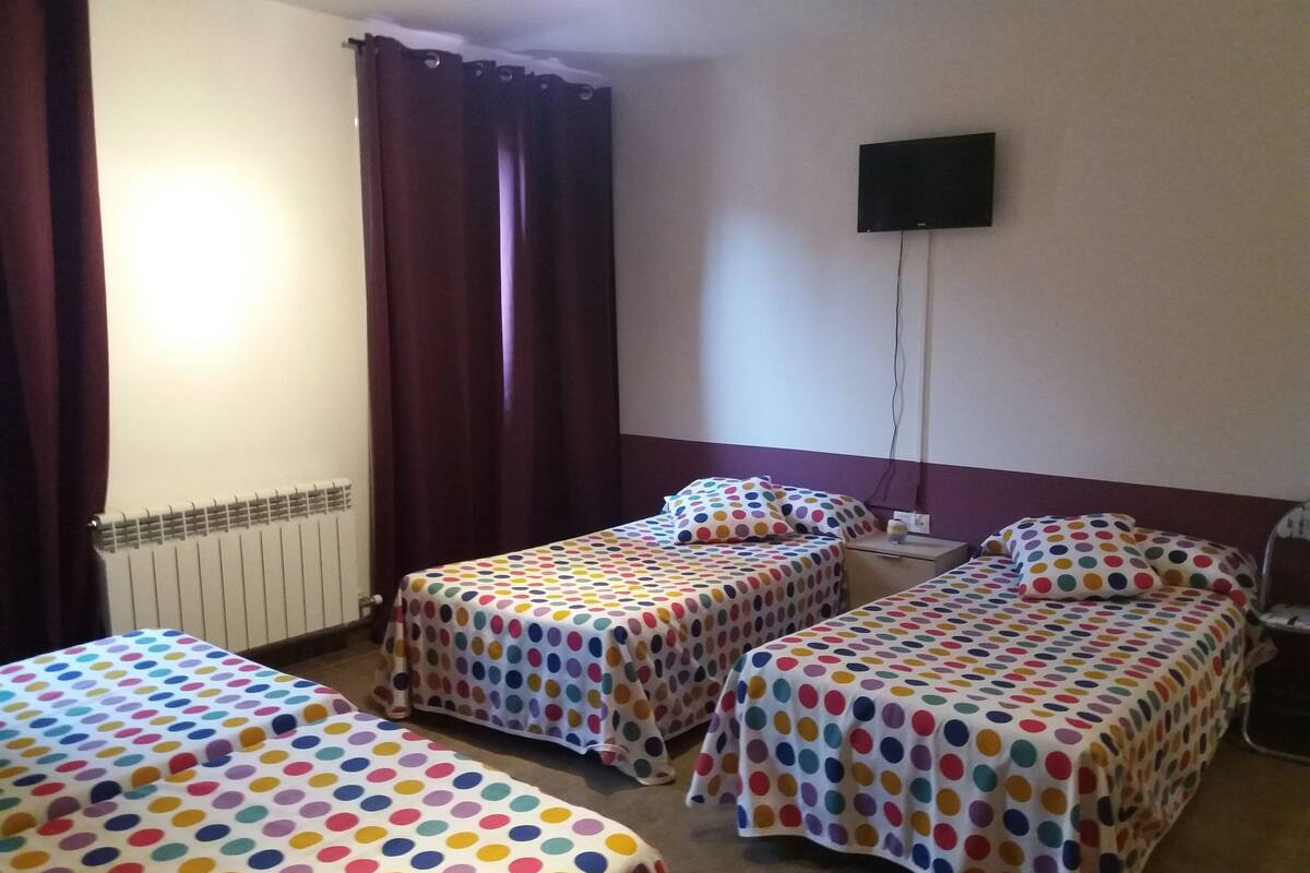 Et hotelværelse med tre enkeltsenge, hver med et pudsetæppe med prikker, et fladskærms-tv monteret på væggen og en radiator.