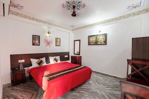 Una habitación de hotel con una cama grande, cabecera de madera y colcha roja. Hay dos mesitas de noche, un espejo y un armario de madera. Las paredes están decoradas con cuadros y un diseño floral en el techo.