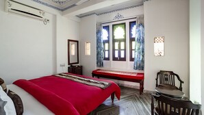 A bedroom with a large bed, a bench, a chair, and a window with curtains. - Janak Niwas (Udaipur)
