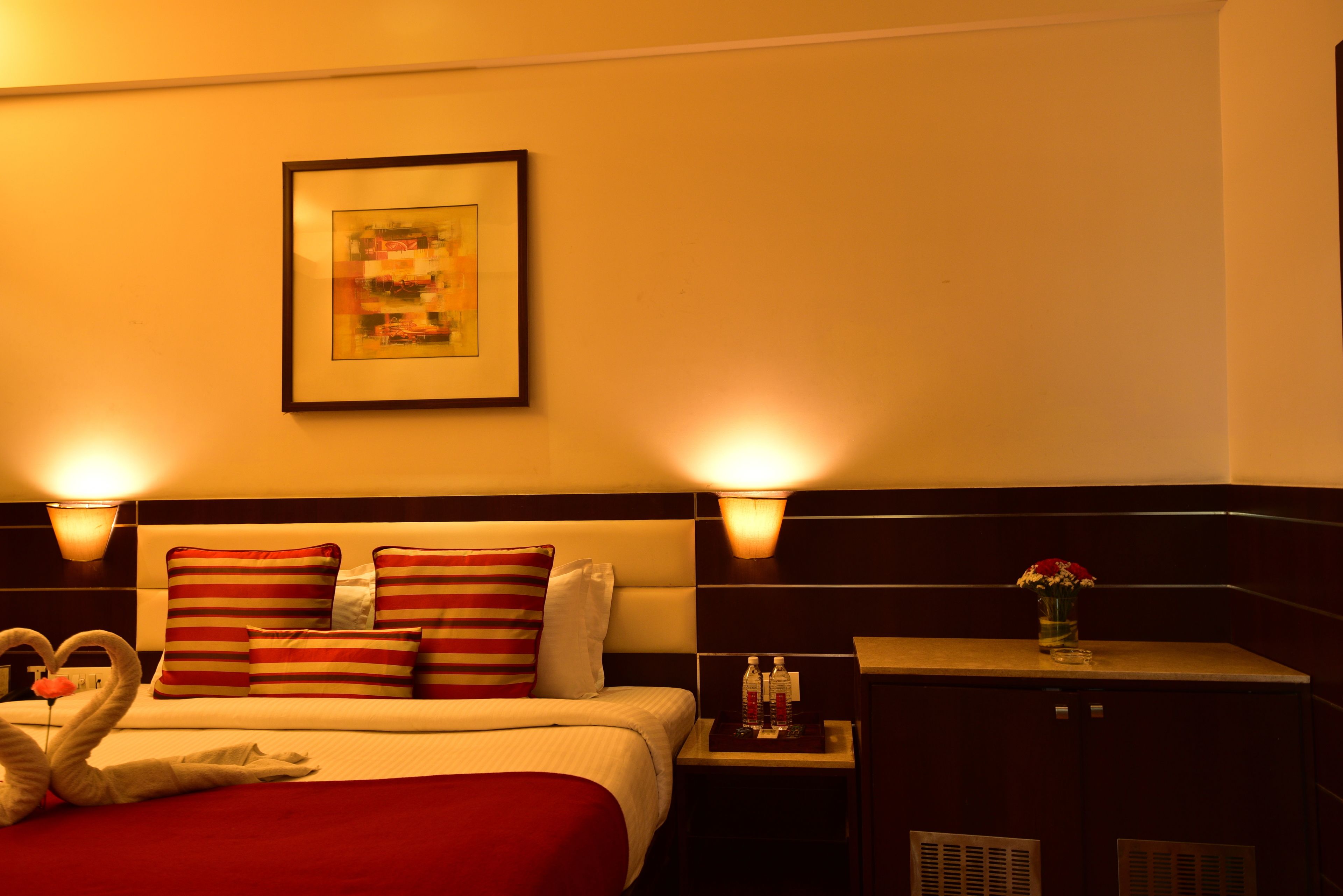 A hotel room with a neatly made bed, striped pillows, a framed picture on the wall, and two wall-mounted lamps.