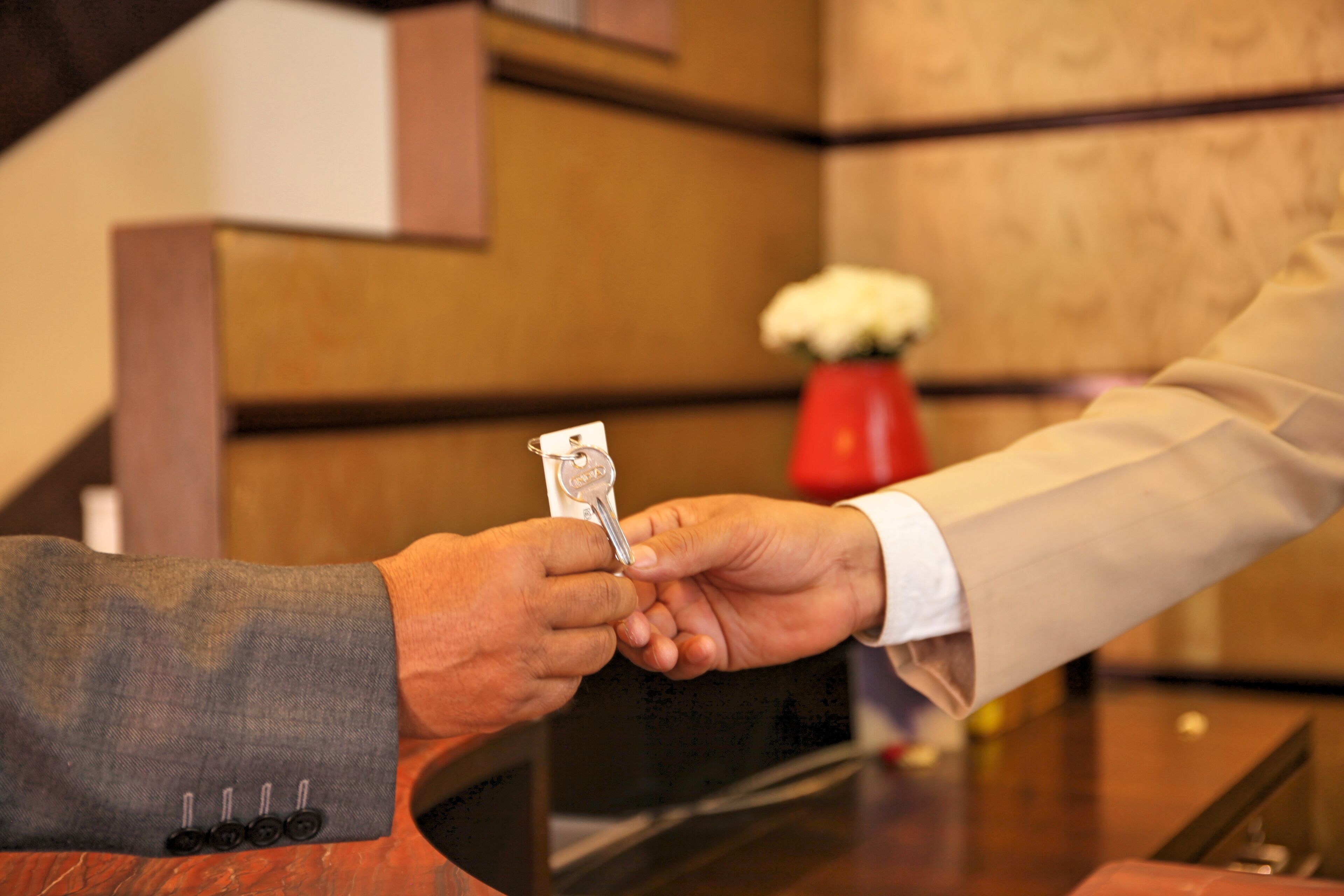 A hand holding a key, another hand receiving it, and a red vase with white flowers in the background.