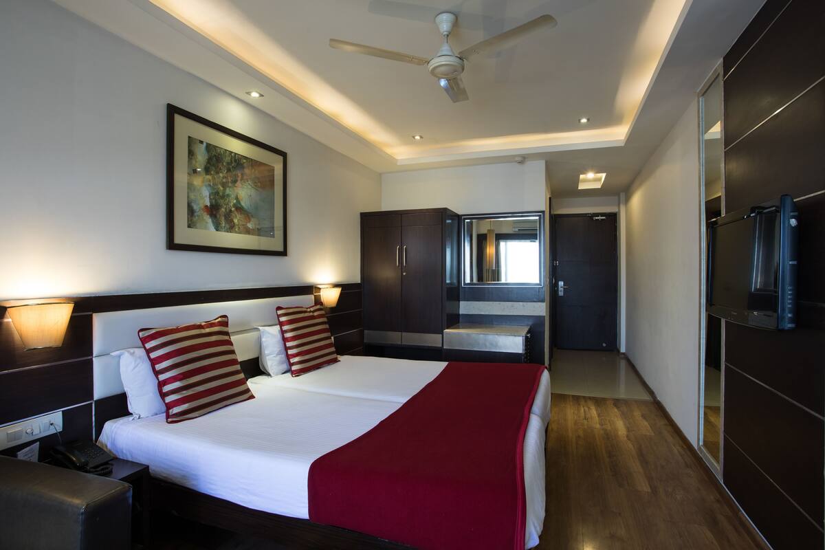 A hotel room with a bed, bedside lamps, a ceiling fan, a painting on the wall, and a view into the bathroom.