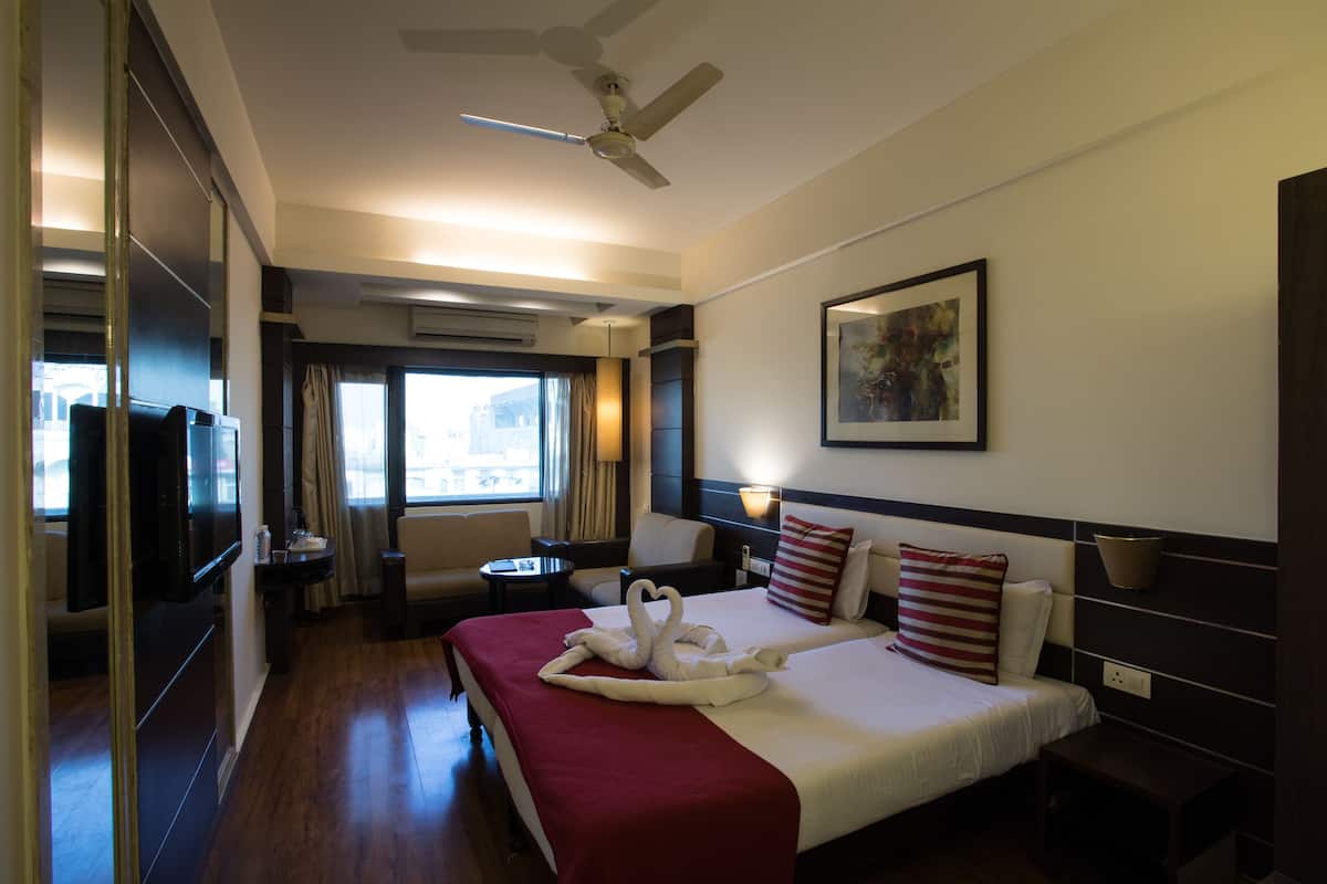 A hotel room with a bed, a TV, a ceiling fan, a window with curtains, and a painting on the wall.